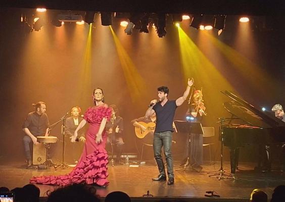 Apresentação musical em um palco com iluminação amarela e alaranjada. Ao centro, uma cantora usa um longo vestido rosa-choque com babados e cauda, enquanto um homem ao lado canta ao microfone com um braço levantado. Ao fundo, músicos tocam violão, violino e percussão. À direita, um pianista toca um piano de cauda preto. A plateia aparece em silhueta na parte inferior da imagem.