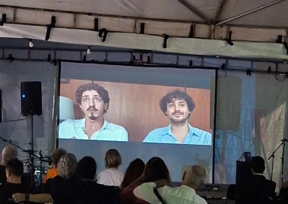 Público sentado em cadeiras assiste a uma sessão de cinema sob uma tenda. Na tela de projeção, aparecem dois homens de cabelos escuros e bigodes, usando camisas azuis claras, diante de um fundo amadeirado. À esquerda, há uma caixa de som; nas laterais, equipamentos e suportes de áudio. O ambiente é pouco iluminado, com a luz da projeção em destaque.
