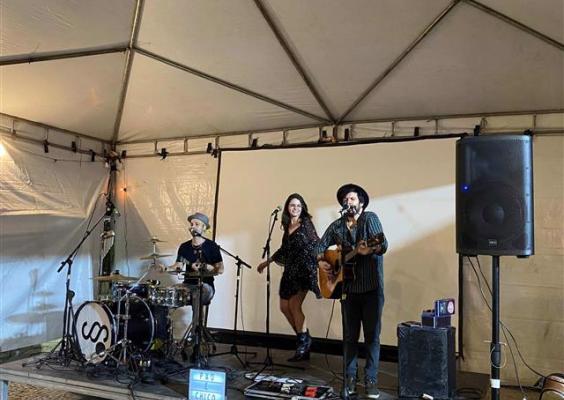 Apresentação musical em um palco montado dentro de uma tenda. Ao centro, uma mulher de vestido preto curto canta ao lado de um homem de chapéu escuro, que toca violão e usa um microfone. À esquerda, um músico toca bateria. Ao fundo, há uma tela branca; nas laterais, caixas de som, microfones, cabos e equipamentos de áudio completam a estrutura do show.