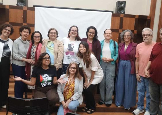Grupo de 13 pessoas posa para uma foto em ambiente interno, diante de uma tela branca e uma parede revestida de madeira. Onze pessoas estão em pé e duas estão agachadas à frente. Vestem roupas casuais de diferentes cores e estilos, e várias usam óculos. Ao fundo, há caixas de som e mobiliário de madeira. A imagem registra um encontro coletivo em um espaço destinado a atividades culturais ou educativas.