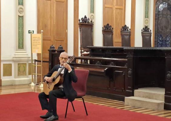 Homem careca, de óculos e vestido de terno preto, toca violão clássico sentado em uma cadeira sobre um tapete vermelho. Ao fundo, destaca-se um mobiliário de madeira escura com cadeiras altas ornamentadas, portas de madeira e paredes decoradas em tons claros e verdes. À direita, há dois degraus de pedra junto à estrutura de madeira. O ambiente é amplo e solene, com piso de madeira listrado.