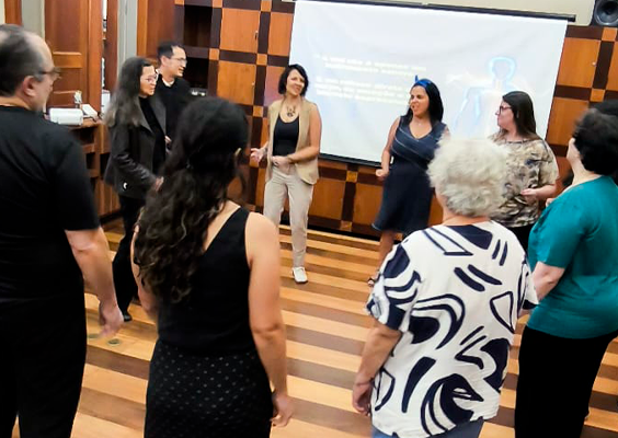 Grupo de pessoas participa de uma atividade em círculo em uma sala com piso de madeira e paredes revestidas de painéis geométricos. Ao fundo, quatro pessoas estão em pé diante de uma tela de projeção, enquanto outras participantes, de diferentes idades, formam o círculo. Elas vestem roupas casuais e parecem interagir durante uma dinâmica coletiva.