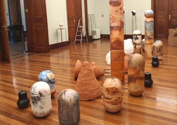 Sala ampla com piso de madeira e portas altas de madeira escura, onde estão expostas diversas esculturas de formas cilíndricas, arredondadas e cônicas, em diferentes tamanhos. As peças apresentam tons terrosos, brancos, azuis e pretos, com texturas e padrões variados. Ao centro, destacam-se esculturas de cerâmica com superfícies rugosas e formas orgânicas.