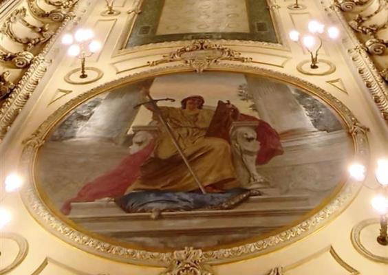 Vista do teto ornamentado de um edifício histórico, com molduras em relevo, detalhes dourados e luminárias acesas. No centro, um grande painel oval pintado retrata uma figura humana com vestes amarelas e vermelhas, segurando uma lança e acompanhada por uma figura animal alongada aos seus pés. A pintura é cercada por uma moldura decorativa, com elementos clássicos e arabescos.