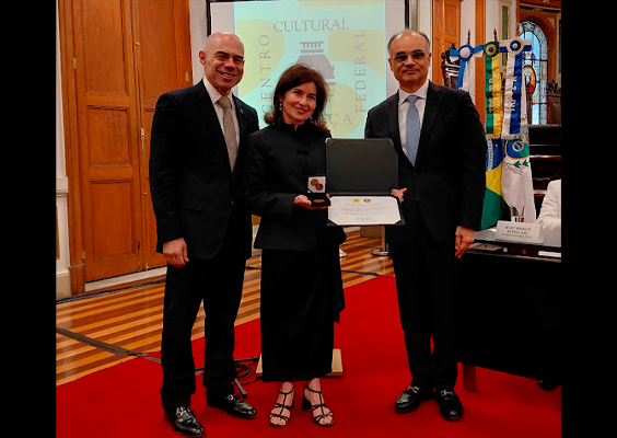 Fotografia de uma cerimônia de homenagem em um salão solene. Ao centro, uma mulher de roupa preta segura uma caixa aberta com uma medalha e um certificado. Ela está entre dois homens de terno escuro, todos sorrindo para a foto. Ao fundo, um homem de terno branco está sentado à mesa. O ambiente tem tapete vermelho, portas de madeira, vitrais, bandeiras e teto com ornamentação detalhada