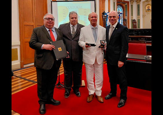 Fotografia de quatro homens em uma cerimônia de homenagem, posando lado a lado sobre um tapete vermelho. O homem à esquerda, de terno escuro e gravata vermelha, segura uma pasta preta. O terceiro, de terno branco, segura uma pequena caixa com medalha, assim como o homem à direita, de terno azul-marinho. Ao fundo, há portas de madeira, bandeiras, cadeiras e um teto ricamente ornamentado