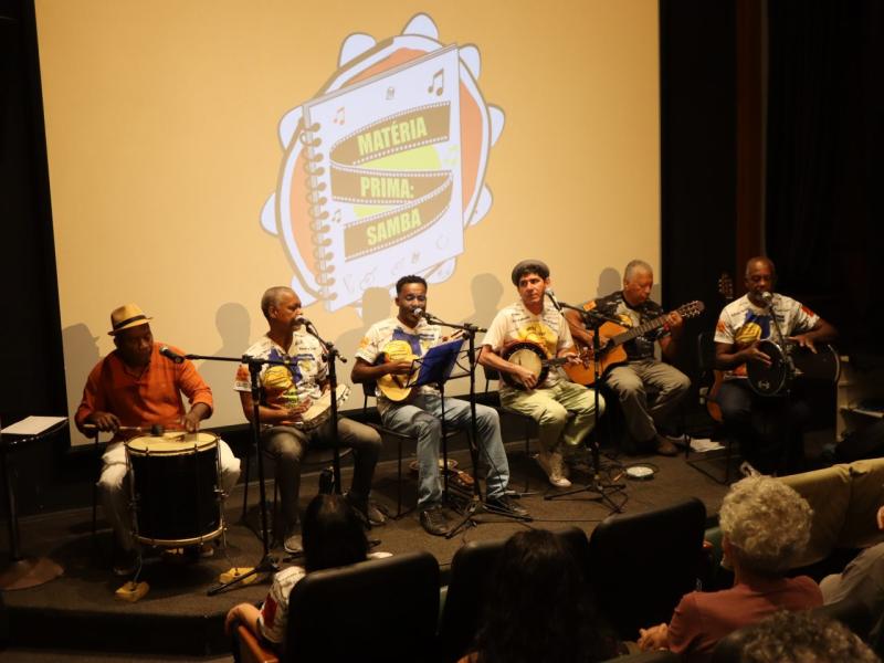 Um grupo de músicos, se apresentam em um palco. Todos estão sentados, tocando e cantando. Entre os instrumentos musicais, estão: pandeiro, banjo, violão, cavaquinho e tan tan.