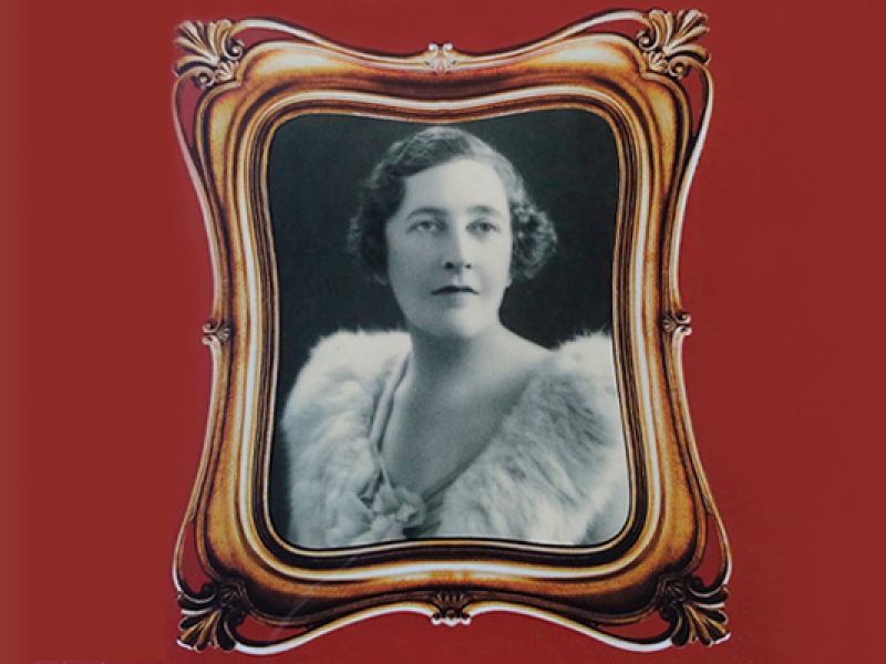 Na imagem, há um porta-retrato de madeira antigo retangular, com arabescos nos cantos. Nele, a foto em preto e branco de época de uma mulher de cabelos curtos (na altura das orelhas), ondulados e castanhos. Ela tem um olhar vago e traços finos. Usa um casaco de pele claro, felpudo. Em volta da moldura, um fundo de cor vermelho queimado.
