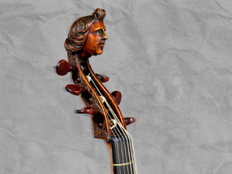 A foto mostra a parte superior (chamada de mão ou cabeça) de um instrumento musical de cordas. É possível observar a ‘mão’ do objeto, toda feita em madeira, com as tarraxas, cordas, trastes e casas. Entalhada no topo, uma cabeça de uma pessoa de cabelos na altura do pescoço, sorrindo levemente, semelhante ao estilo antigo, de época. O plano de fundo é cinza claro, com textura.