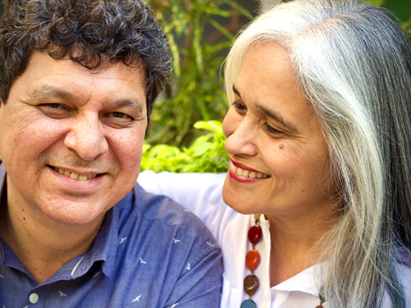 Na foto, close-up de duas pessoas, um homem e uma mulher brancos. Ele tem cabelos enrolados castanhos e sorri para a câmera; usa camisa de botões azul com o desenho de pequenas gaivotas brancas. Ela olha para ele sorrindo, está mais de perfil; usa batom vermelho, blusa branca, colar colorido e tem cabelos grisalhos com uma grande mecha branca emoldurando o rosto. O plano de fundo é formado por arbustos e folhagens.