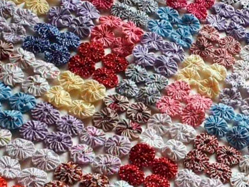 A foto mostra parte de uma toalha (ou lençol) de mesa feito de fuxico (técnica artesanal em que há o reaproveitamento de retalhos de tecido) de cores variadas: vermelho, azul, rosa, amarelo, marrom, lilás etc. Há uma lógica formada por um quadrado de quatro fuxicos de cada cor, um costurado no outro.