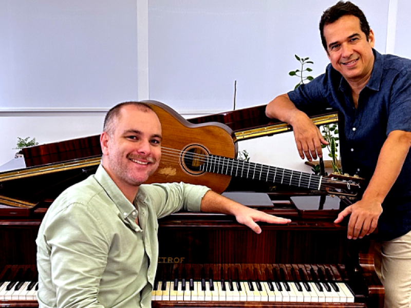 A foto mostra dois homens brancos, um sentado à frente de um piano de cauda e o outro em pé, do lado direito, apoiado no piano. Ambos sorriem para a câmera. O primeiro é grisalho, um pouco calvo, e usa barba cerrada; veste blusa social verde clara. Já o tem cabelos curtos castanhos escuros e usa blusa de botão azul marinho e calça clara. Em cima do piano, de madeira escura envernizada, um violão.