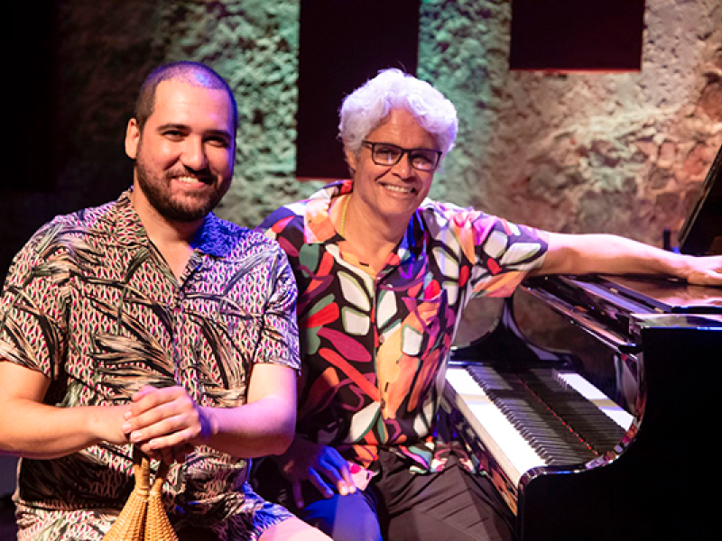 Duas pessoas sorrindo, sentadas ao lado de um piano. Da esquerda para a direita: homem com barba e cabelos curtos escuros segura instrumento de percussão. Ele usa camisa de manga curta com estampas geométricas; homem mais velho, de cabelos brancos, óculos e camisa estampada com cores vibrantes. Ele está sentado ao piano, com a mão esquerda repousando sobre o instrumento. Ao fundo, uma parede de pedra com iluminação suave, sugerindo que estão em um local de apresentação musical, como um palco ou estúdio.