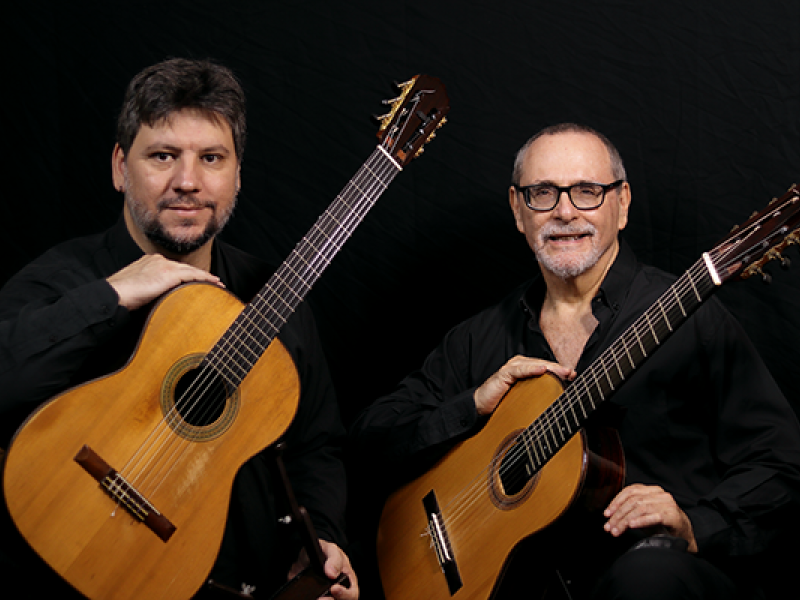 A imagem mostra dois homens sentados, cada um segurando um violão clássico. Ambos vestem camisas pretas e estão de frente para a câmera, com expressões serenas. O homem à esquerda tem barba curta e cabelo castanho, enquanto o da direita usa óculos, tem barba grisalha e está sorrindo. O fundo é preto, destacando os músicos e seus violões. A imagem transmite um clima de seriedade e paixão pela música.