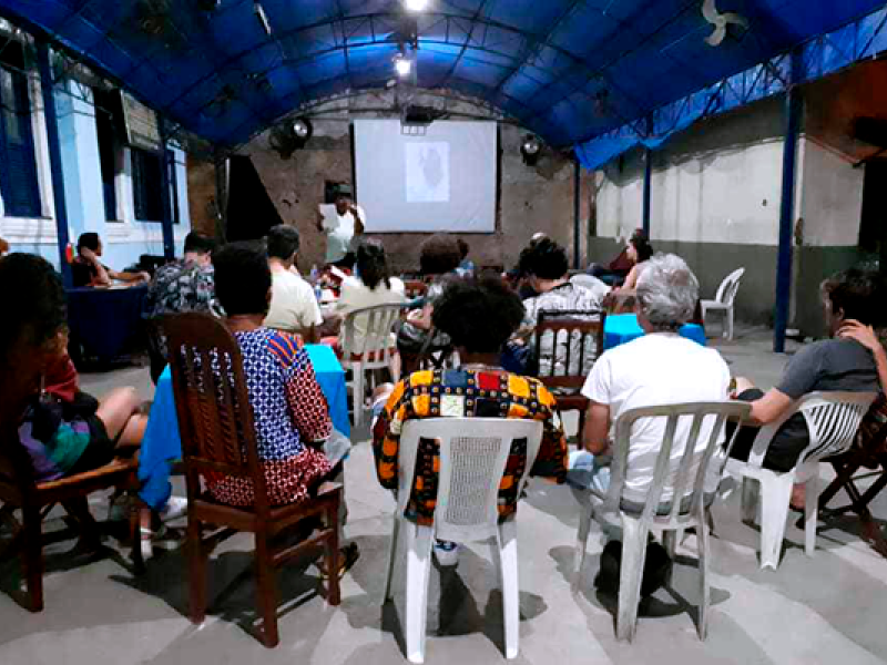 A imagem mostra várias pessoas sentadas em cadeiras de diferentes tipos, olhando para uma tela de projeção em um ambiente coberto, com um teto azul. A sala parece ser um espaço simples e comunitário, com paredes parcialmente desgastadas. Na tela, há uma apresentação, mas o conteúdo não está claro. A iluminação é suave, com foco na tela, e o ambiente sugere uma reunião ou evento informativo.
