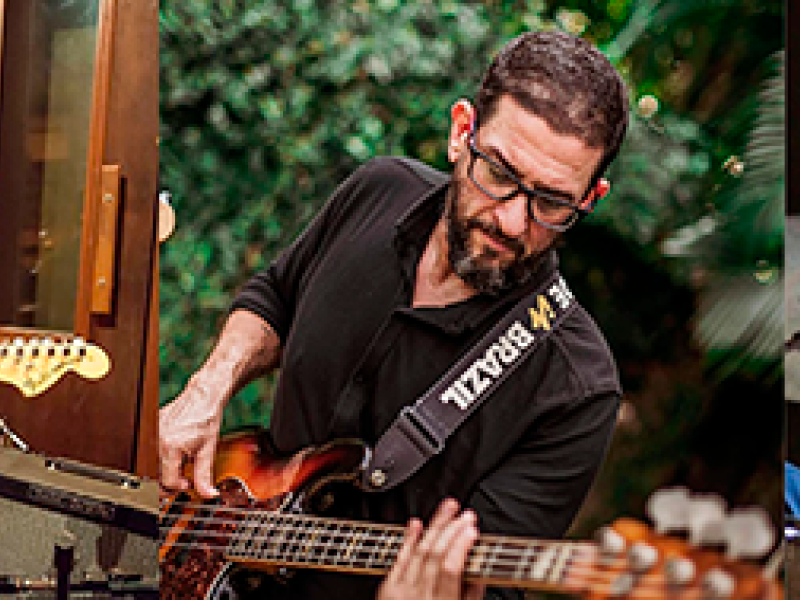 Montagem de três fotos com um musicista cada. Da esquerda para a direita: homem branco de cabelos curtos e escuros (usa blusa preta com mangas curtas e calça clara) toca guitarra em estúdio e sorri para alguém que não aparece na foto; homem branco, barbudo, de cabelos curtos e escuros (veste blusa preta de mangas longas e usa óculos) toca guitarra com o corpo inclinado para frente. Ao fundo, um espaço com plantas; homem negro (usa chapéu, óculos de grau e blusa de manga custa preta) toca bateria.