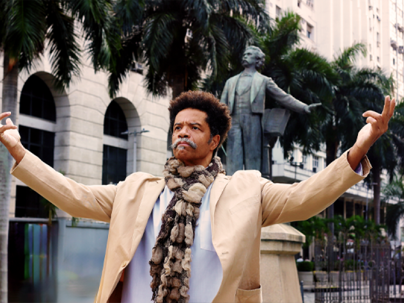 A foto mostra um homem negro de bigode grisalho e cabelos afro com os braços erguidos. Usa blazer bege claro, blusa branca e cachecol em tons marrons. Ele está com um olhar obstinado, à frente de uma estátua de outro homem de bigode e paletó. Há palmeiras (árvores) e prédios no cenário.