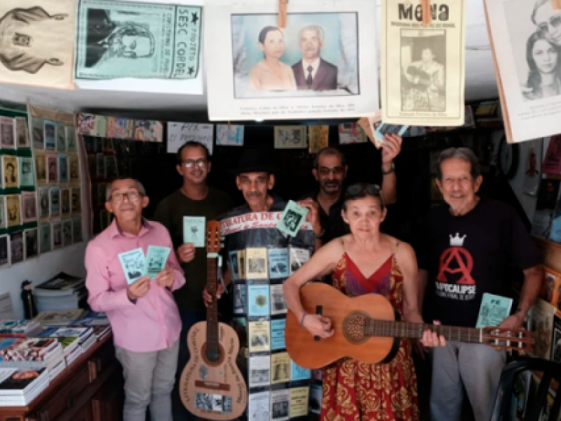 Grupo com 5 homens e uma mulher brancos, em sala com livretos de literatura de cordel. Um deles veste traje plástico transparente com livretos amarelos, azuis e brancos, de cordel. Usa chapéu preto e tem um violão em uma das mãos. Uma mulher de óculos, ao centro, toca outro violão e veste vestido estampado na cor vinho. Os outros homens seguram livretos de cordel, alguns usam óculos e alguns possuem cabelos grisalhos curtos. No 1º plano, na parte superior da imagem, há 5 livros do tema pendurados em varal.