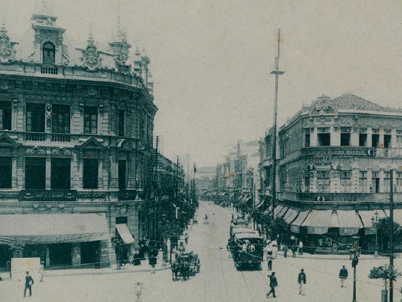 Fotografia antiga do Largo da Carioca, Rio de Janeiro. Os edifícios em destaque possuem arquitetura elaborada com ornamentos detalhados nas fachadas, característica do estilo europeu do início do século XX. A rua é movimentada, com pessoas caminhando e alguns veículos de tração animal. Há também uma linha de bonde no centro da via. O cenário transmite uma atmosfera clássica e urbana, com foco na vida cotidiana da época.