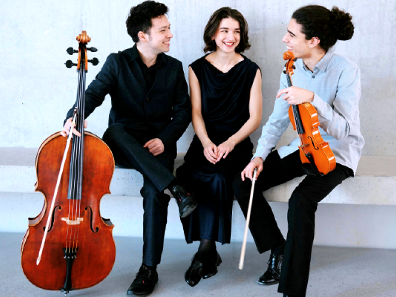 Na foto, dois homens e uma mulher branca estão sentados em um banco; todos sorriem. Um deles (à esquerda) usa paletó preto e segura um violoncelo. A mulher, no meio dos dois, veste vestido longo preto e olha para um dos musicistas. O homem da direita usa blusa social clara e calça preta; ele segura um violino.