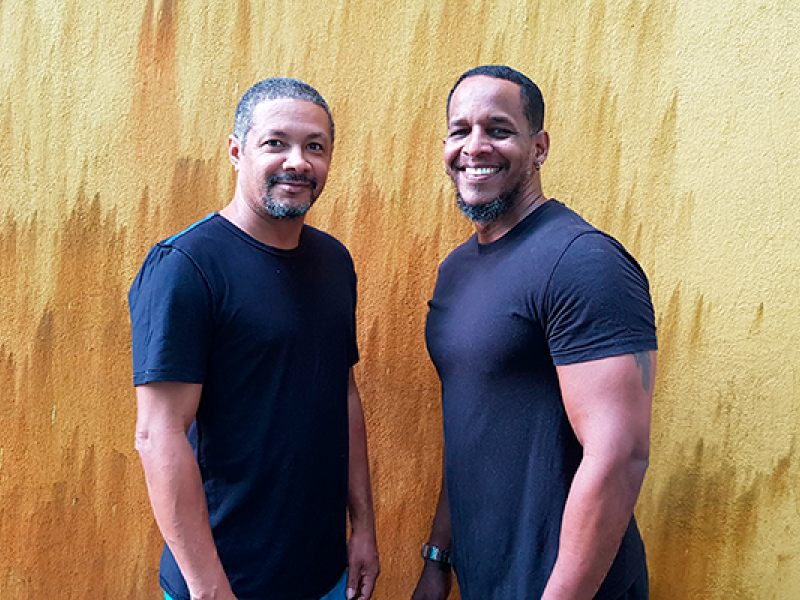 A imagem mostra dois homens posando juntos em frente a uma parede com uma textura amarelada e manchas de cor mais escura. Ambos estão usando camisetas pretas e calças escuras. Eles sorriem, criando uma atmosfera amigável e descontraída. Os dois têm cabelo curto e barba, e parecem confortáveis na foto. A simplicidade do fundo e das roupas realça o foco nos rostos e expressões dos homens.