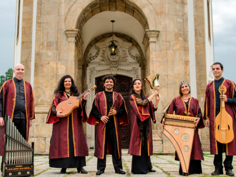 A imagem mostra um grupo de seis músicos, todos vestidos com trajes tradicionais, longos e em tons de vinho com detalhes dourados. Eles estão posando em frente a um edifício antigo, que parece ser uma igreja com uma arquitetura clássica de pedra. Cada um está segurando um instrumento musical antigo: alaúdes, flauta, um órgão portátil, uma harpa e um par de sinos. O grupo aparenta ser especializado em música antiga ou medieval, e o cenário reforça essa atmosfera histórica e cultural.