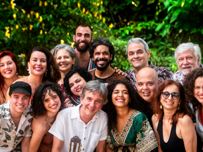 Na foto, um grupo de 15 pessoas, entre homens e mulheres das mais variadas faixas etárias, sorri para a câmera. Eles estão em uma área aberta, com vegetação ao fundo. Há mulheres de cabelos castanhos lisos e enrolados, ruiva, e grisalhas; homens de cabelos castanhos curtos, castanhos enrolados, careca e grisalhos. As roupas se misturam entre estampadas e lisas (brancas e pretas). É possível perceber a ponta de um violão na mão de um dos homens que está posicionado ao centro, na fileira da frente da foto.