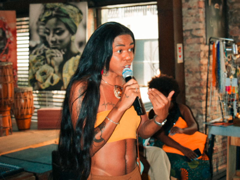 A imagem mostra uma mulher negra de cabelo comprido, liso e escuro, usando um top amarelo e segurando um microfone enquanto fala. Ela tem uma tatuagem visível no braço esquerdo e acessórios nos braços e no pescoço. Ao fundo, há uma decoração rústica com tambores e quadros, incluindo um grande retrato de uma mulher com um turbante. A atmosfera parece ser a de um evento cultural ou artístico em um espaço acolhedor, com outra mulher negra sentada no fundo.