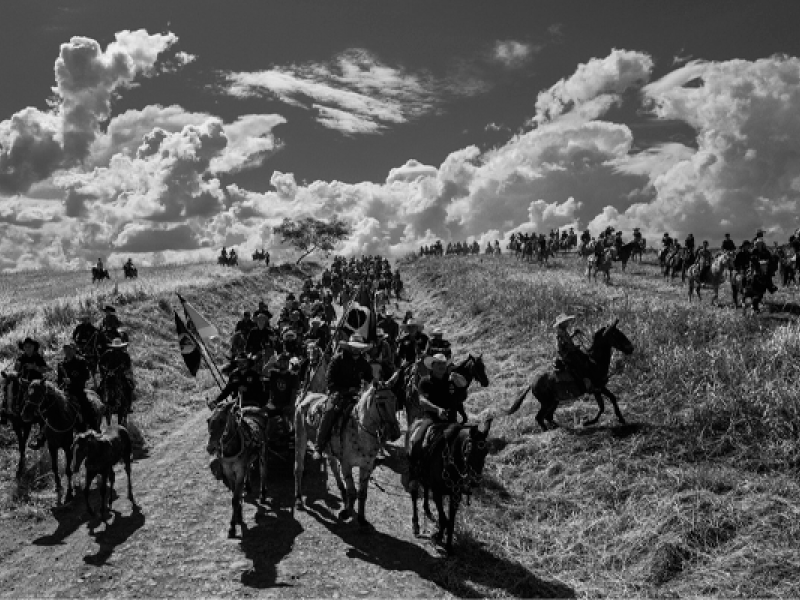 A imagem mostra um grande grupo de cavaleiros em uma paisagem rural. Os cavaleiros estão organizados em fileiras, cavalgando por uma estrada de terra com bandeiras do Brasil ao vento. O cenário é dominado por um céu cheio de nuvens dramáticas. Ao fundo, há mais cavaleiros espalhados pelo campo, criando uma sensação de movimento e intensidade. A imagem é em preto e branco, destacando os contrastes de luz e sombra, enquanto a vasta planície se estende ao redor.