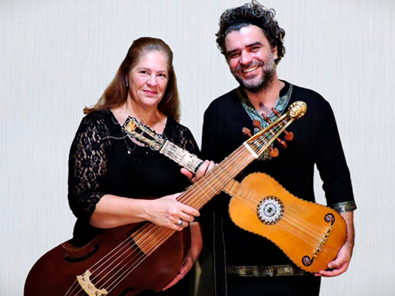 A imagem mostra duas pessoas, uma mulher e um homem, sorrindo enquanto seguram instrumentos musicais antigos. A mulher, com cabelos longos e claros, veste uma blusa preta rendada e segura um instrumento de cordas grande, semelhante a uma viola da gamba. O homem, de cabelos escuros e barba, usa uma camisa preta com detalhes bordados e segura um instrumento de cordas menor, que se assemelha a uma guitarra barroca. Ambos parecem orgulhosos.