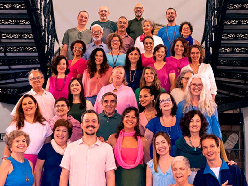 A imagem mostra um grande grupo de pessoas posando na escadaria do CCJF, formando várias fileiras. Eles estão vestidos com roupas coloridas, predominantemente em tons de rosa, azul, verde e roxo, e todos parecem sorrir para a câmera, transmitindo uma atmosfera alegre e amistosa. As idades dos participantes variam, incluindo adultos de meia-idade e idosos.