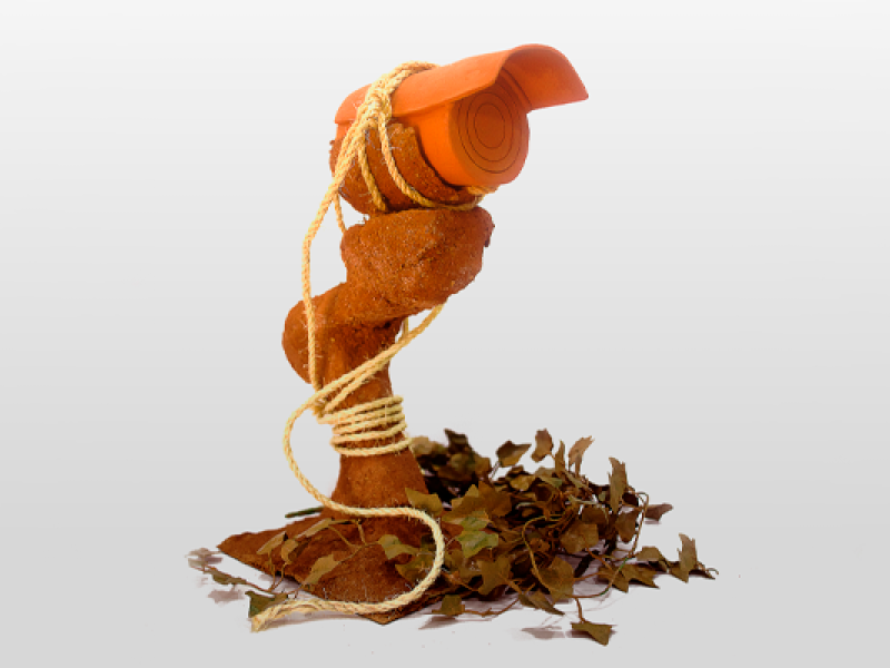 A imagem mostra um objeto semelhante a uma câmera de vigilância que parece ser feita de E.V.A laranja enrolado e amarrado, por uma corda, a um tronco ou um objeto rústico de barro. Há folhas secas espalhadas na base, criando um ambiente natural. O objeto parece representar uma fusão de tecnologia (câmera) com elementos da natureza, sugerindo um contraste entre o artificial e o orgânico.