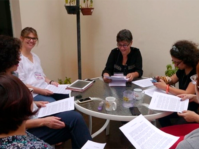 A imagem mostra um grupo de sete pessoas sentadas ao redor de uma mesa redonda de vidro, em um ambiente interno iluminado. Elas estão segurando papéis e parecem envolvidas em uma discussão ou reunião. A pessoa ao centro, de cabelos curtos e grisalhos, está sorrindo e segurando documentos, enquanto outras estão lendo ou fazendo anotações. Na mesa, há xícaras de café, um tablet e outros objetos pessoais. O ambiente sugere uma reunião colaborativa, possivelmente de trabalho ou estudo.