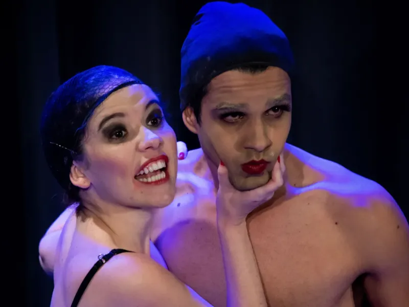 A imagem mostra dois atores em um cenário teatral com iluminação dramática. A mulher, à esquerda, tem expressão exagerada e está sorrindo de forma teatral, segurando o rosto do homem com uma das mãos. Ambos usam maquiagem marcante, com batom vermelho e sombra preta destacando os olhos. O homem, à direita, está com uma expressão séria e levemente melancólica. Ambos usam toucas pretas, e a iluminação azul e violeta acentua o tom performático e estilizado da cena.