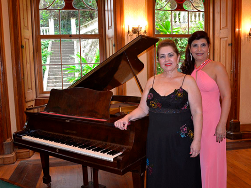 A imagem mostra duas mulheres posando ao lado de um piano de cauda em uma sala elegante com decoração clássica. A mulher à esquerda usa um vestido preto com alças finas e bordados coloridos, enquanto a da direita veste um vestido longo rosa claro com detalhes brilhantes. Ao fundo, há janelas grandes que mostram um jardim com escadas e vegetação. O ambiente é iluminado por luz natural e candelabros nas paredes, sugerindo um espaço refinado e aconchegante.