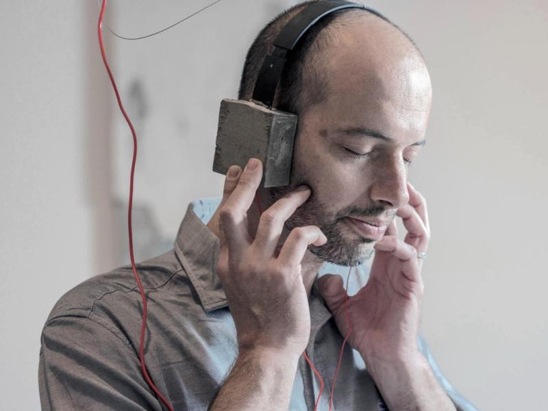 A imagem mostra um homem calvo, de pele clara, usando um fone de ouvido grande enquanto segura um pequeno dispositivo próximo à orelha. Ele parece concentrado, com a outra mão apoiando o rosto, sugerindo que está ouvindo ou refletindo atentamente. O ambiente ao fundo é claro, simples e levemente desfocado, destacando o homem como o foco principal da composição.