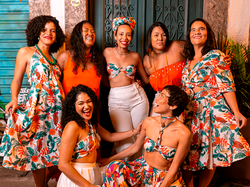 A imagem mostra um grupo de sete mulheres sorrindo e posando juntas em frente a uma porta, em ambiente externo. Elas usam roupas coloridas com estampas tropicais e vibrantes, predominando tons de laranja, verde e branco. Alguns vestem vestidos, enquanto outros usam tops e saias, criando uma estética alegre e descontraída. O cenário urbano e a expressão das mulheres passam uma sensação de celebração e camaradagem.