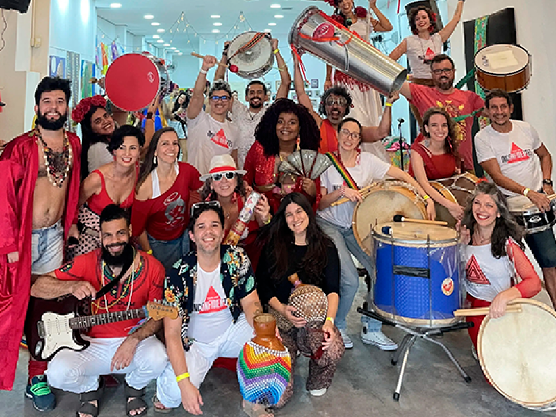 A imagem mostra um grupo alegre de pessoas em um espaço interno, segurando instrumentos de percussão como surdos, tamborins e chocalhos. Algumas vestem fantasias e roupas coloridas, criando um clima festivo. Todos sorriem e posam para a foto, transmitindo um ambiente de celebração, possivelmente de uma escola de samba ou grupo de música brasileira. Ao fundo, a decoração é simples e colorida, reforçando a atmosfera vibrante