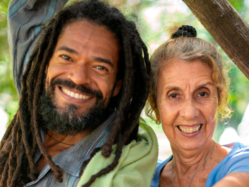 A imagem mostra duas pessoas sorrindo para a câmera em um ambiente ao ar livre, iluminado por luz natural. À esquerda, há um homem com cabelos longos e estilo dreadlocks, barba cheia e um sorriso caloroso. Ele veste uma camisa e uma peça verde jogada sobre os ombros. À direita, há uma mulher de cabelos claros presos em um coque, também sorrindo, transmitindo simpatia. O fundo desfocado sugere um cenário natural, com árvores ou vegetação. A foto passa uma sensação de alegria e conexão entre as pessoas.