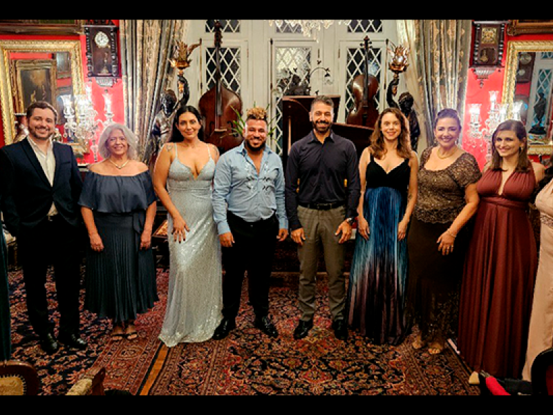 Grupo de pessoas posando em um ambiente elegante e decorado de forma clássica, com paredes vermelhas, lustres, quadros e mobília detalhada. Todos estão vestidos formalmente, com trajes de gala como vestidos longos e ternos, sugerindo que estão participando de um evento especial ou cerimônia. O ambiente tem um tapete com padrão floral e móveis sofisticados, criando uma atmosfera luxuosa e histórica. Ao fundo, há detalhes arquitetônicos, como janelas em estilo gótico e estatuetas decorativas