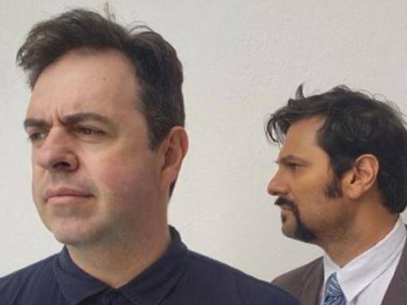 A imagem mostra dois homens em plano médio. O homem à frente tem cabelos castanhos curtos, veste uma camisa azul-escura e olha para a esquerda. O homem ao fundo, ligeiramente desfocado, tem cabelos castanhos escuros, barba curta e veste uma camisa azul clara com gravata listrada, acompanhado de um blazer cinza. Ambos estão posicionados contra um fundo branco liso.