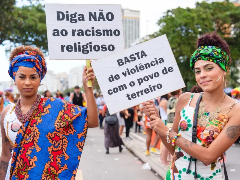 A imagem mostra duas mulheres segurando cartazes em um evento ao ar livre. A da esquerda usa um turbante azul com estampa colorida e um vestido combinando. A da direita usa um turbante verde estampado e roupas com detalhes brancos e verdes. O cartaz da esquerda diz: "Diga NÃO ao racismo religioso", e o da direita: "BASTA de violência com o povo de terreiro". Elas estão em uma manifestação, possivelmente relacionada à valorização de religiões afro-brasileiras.