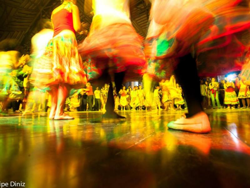 A imagem mostra uma cena dinâmica de dança em grupo, com pessoas girando em um salão iluminado por luzes amarelas e coloridas, criando um efeito de movimento borrado. Os dançarinos vestem roupas tradicionais, como saias rodadas e coloridas, típicas de danças folclóricas brasileiras. O reflexo das cores no piso contribui para uma atmosfera vibrante e festiva. A foto, assinada por Felipe Diniz, capta a energia e a vivacidade da dança popular.