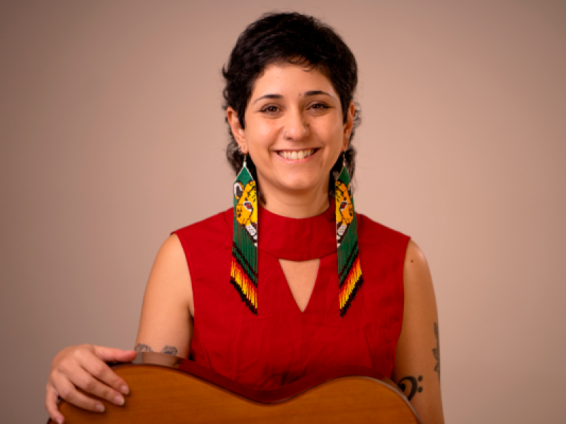 A imagem retrata uma mulher sorridente em um fundo neutro. Ela usa um vestido vermelho e ostenta longos brincos artesanais com motivos indígenas, destacando cores vibrantes como amarelo, verde e preto, e figuras de onças. Seu cabelo é curto e ondulado, e ela possui uma tatuagem visível no braço esquerdo e um piercing no nariz. A mulher está segurando um violão, sugerindo uma conexão com a música ou a arte. A expressão dela transmite simpatia e confiança.