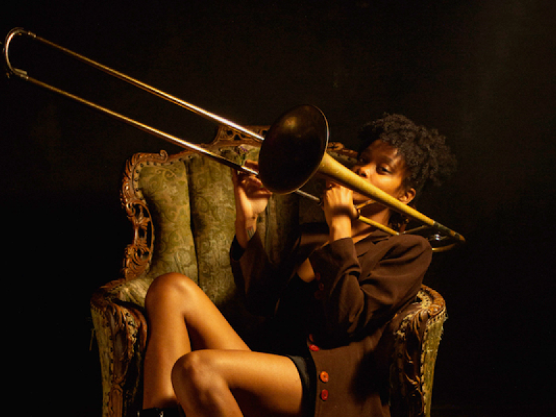 A imagem mostra uma mulher tocando trombone, sentada em uma poltrona vintage de estilo clássico. A cena possui uma iluminação dramática, com destaque para a musicista e seu instrumento, que são iluminados em contraste com o fundo escuro. A postura e expressão da artista, aliadas ao ambiente intimista, sugerem uma atmosfera de concentração e paixão pela música. O trombone e a poltrona de design antigo reforçam a conexão com um ambiente sofisticado ou artístico.