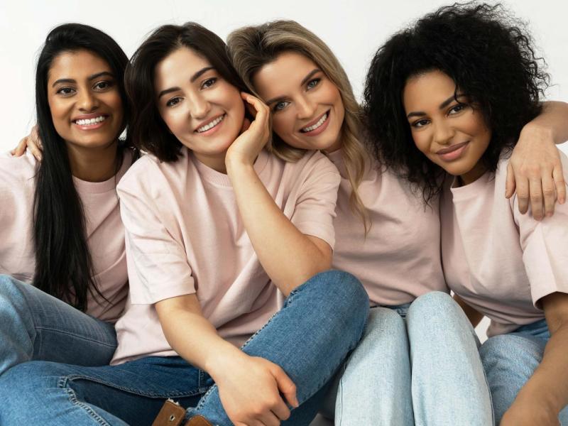 A imagem mostra quatro mulheres sentadas juntas, vestindo camisetas rosa claro e calças jeans. Elas estão sorrindo e abraçadas, transmitindo um forte senso de amizade e união. Cada uma possui traços e estilos distintos, representando diversidade. O fundo é branco, destacando o grupo. A imagem evoca sentimentos de apoio, companheirismo e felicidade.