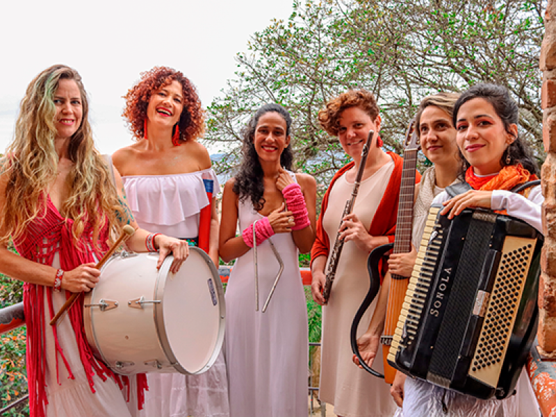 A imagem mostra seis mulheres sorridentes em uma sacada, com árvores ao fundo e uma estrutura de tijolos na lateral. Elas vestem roupas predominantemente brancas com detalhes em vermelho e seguram instrumentos musicais (tambor, triângulo, flauta, violão e sanfona). A cena transmite uma atmosfera leve e artística, sugerindo que fazem parte de um grupo musical. O sorriso e a postura das mulheres indicam alegria e cumplicidade.