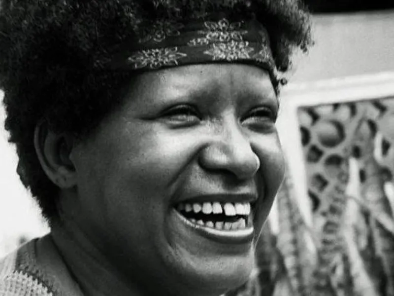A imagem em preto e branco mostra Lélia Gonzalez sorrindo amplamente. Seu rosto expressa alegria, com olhos levemente semicerrados e dentes à mostra. Seu cabelo crespo é adornado por uma faixa estampada na testa. O fundo apresenta elementos arquitetônicos vazados e algumas plantas, sugerindo um ambiente externo ou uma varanda. A luz suave realça os contornos do rosto, destacando sua expressão vibrante e acolhedora.