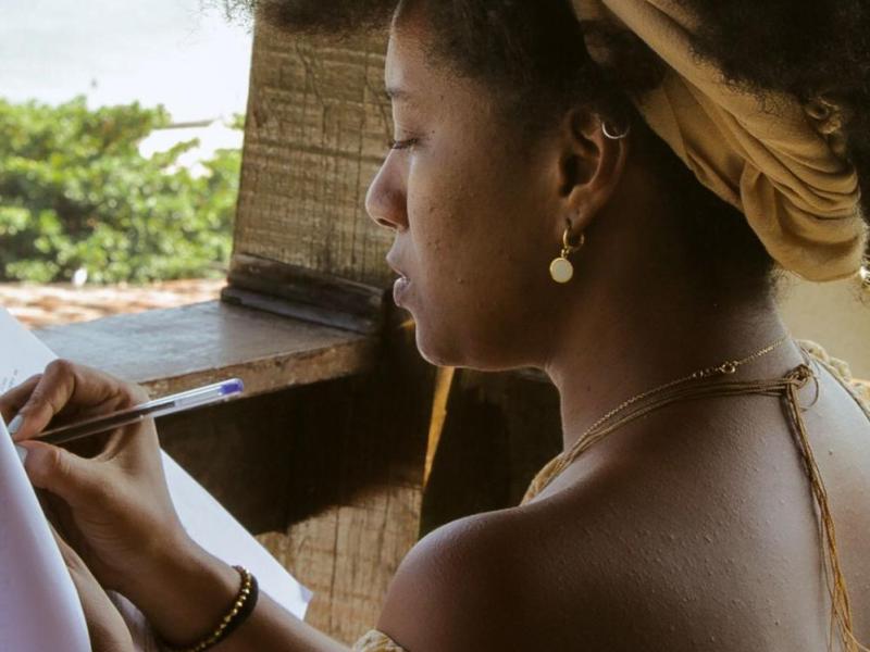A imagem mostra uma mulher negra de perfil, escrevendo ou desenhando em um papel com uma caneta azul. Ela usa um turbante bege na cabeça, brincos dourados e colares finos. Seu braço está adornado com pulseiras. O ambiente tem uma janela de madeira aberta, com vista para árvores, sugerindo um local ao ar livre ou próximo à natureza. A luz natural ilumina seu rosto, destacando sua expressão concentrada.