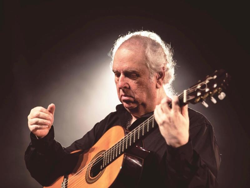 A imagem mostra o violonista Edelton Gloeden em um momento de concentração enquanto toca seu violão. Com expressão serena e gestos precisos, ele veste uma camisa preta e está envolto por uma iluminação suave que destaca sua presença e cria um clima intimista. A cena transmite a sensibilidade e a profunda conexão de Gloeden com a música erudita e o violão.